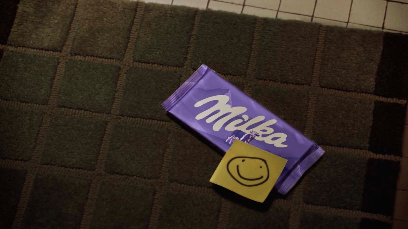 Vor der Wohnungstür des älteren Mannes liegt auf der Fussmatte eine Tafel Milka Alpenmilch Schokolade mit einem Post-it, der einen Smiley zeigt.